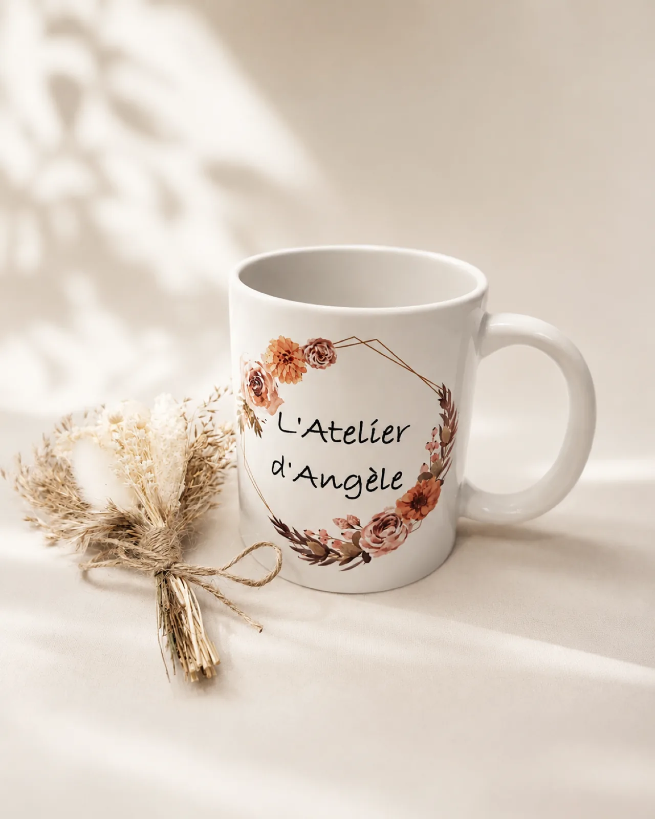 Mug blanc « L’Atelier d’Angèle » orné d’une couronne de fleurs, posé près d’un bouquet de fleurs séchées